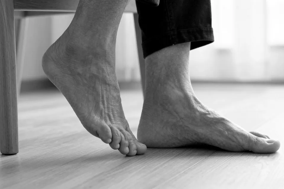 An older adult’s bare feet on a wooden floor beside a chair, one foot flat and the other with the heel raised, in black and white