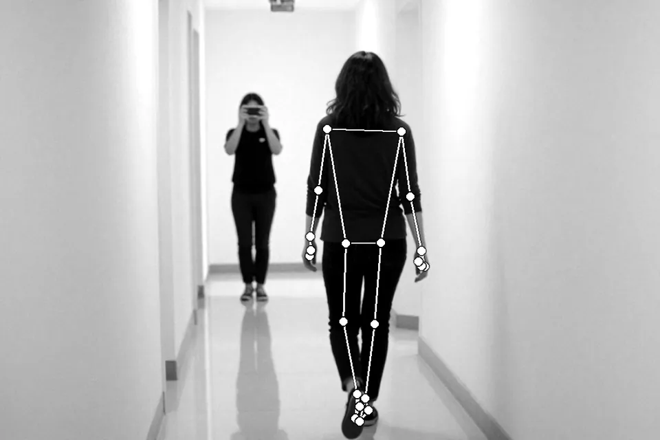 A woman walking down a hallway while a person at the far end films her with a smartphone, with pose-estimation joint lines drawn over the walker, in black and white
