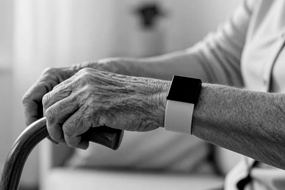 An older woman’s hands resting on the handle of a walking cane, with a small sensor worn on her wrist, in black and white